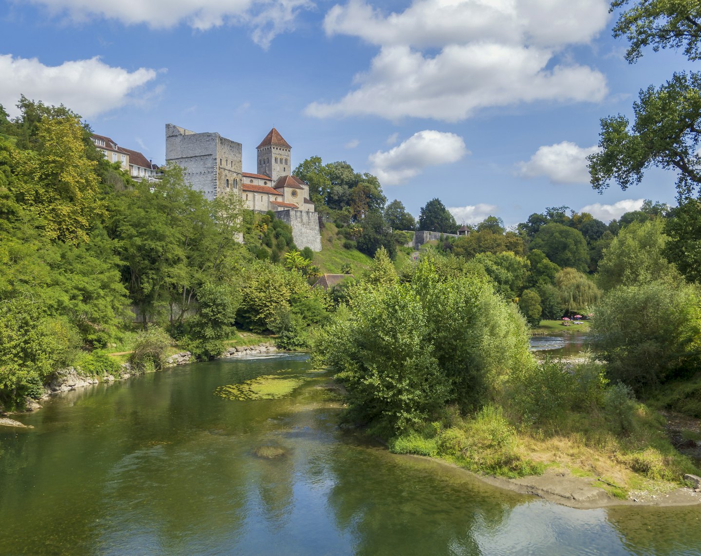 Steinschloss mit roten Dächern auf einem grünen Hügel mit Blick auf einen Fluss.